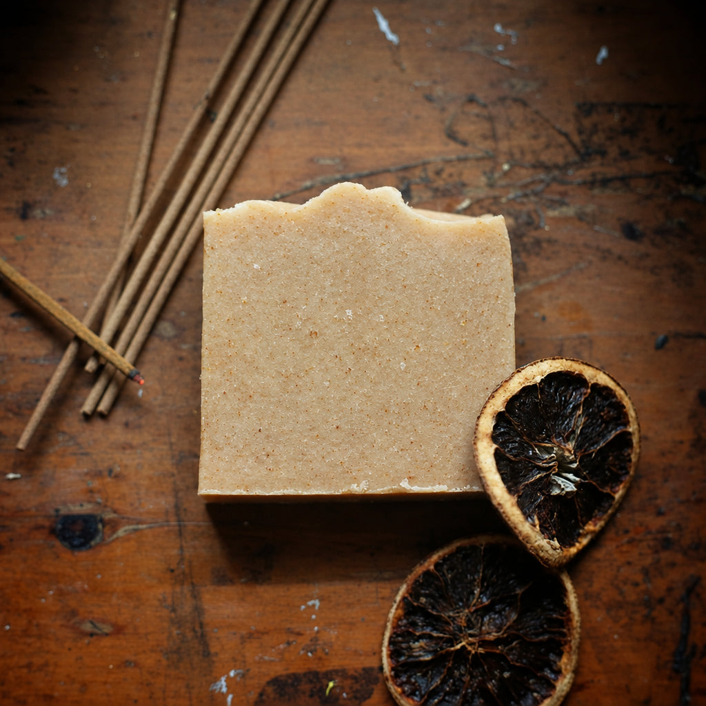 A rectangular bar of pale beige handmade soap sits on a rustic wooden surface, styled with dried dark citrus slices and several thin wooden sticks.
