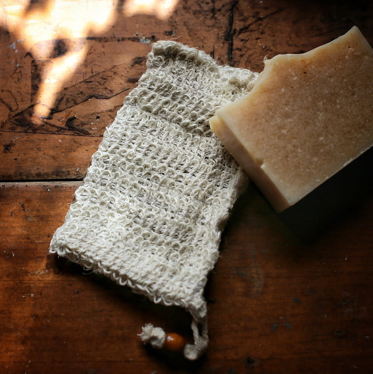 A handmade rectangular bar of soap in a dark container next to a sisal soap bag with a drawstring, set on a rustic dark wooden surface.