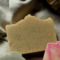 One artisanal handcrafted soap bar with scalloped edges, arranged on a draped beige linen fabric with a piece of dark wood in the background. The focal bar is light tan, laying above one is dusty blue green with pink swirls, and one is pink and cream marbled.