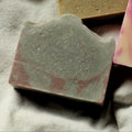 A rectangular handmade soap bar with pink and Indigo swirls, resting on a textured off-white linen cloth next to a gold textured soap and a pink and cream textured soap.