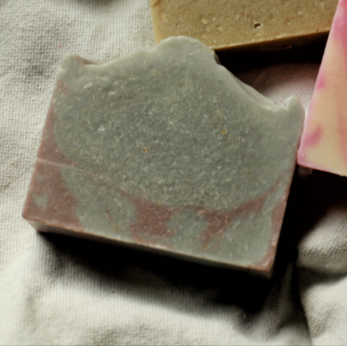 A rectangular handmade soap bar with pink and Indigo swirls, resting on a textured off-white linen cloth next to a gold textured soap and a pink and cream textured soap.