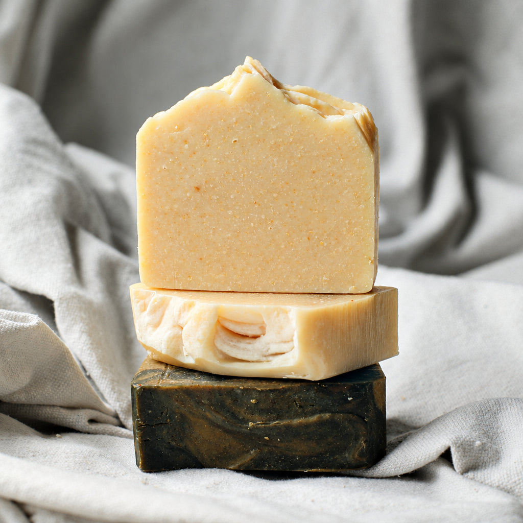 Two stacked bars of handmade soap on a dark wooden block, set against a draped light gray linen fabric background.
