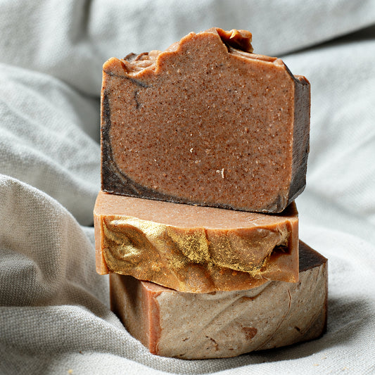 Three stacked handmade soap bars in earthy brown, tan, and kraft paper tones, photographed on draped light fabric.