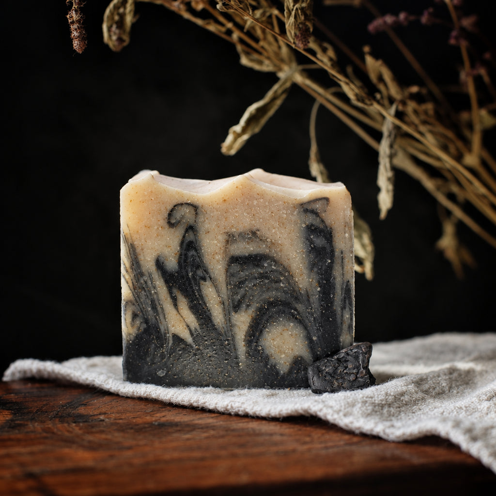 A handmade rectangular bar soap with a swirled black, gray, and cream pattern, resting on a white towel over a wooden surface with dramatic sunlight and dried flowers in the background.