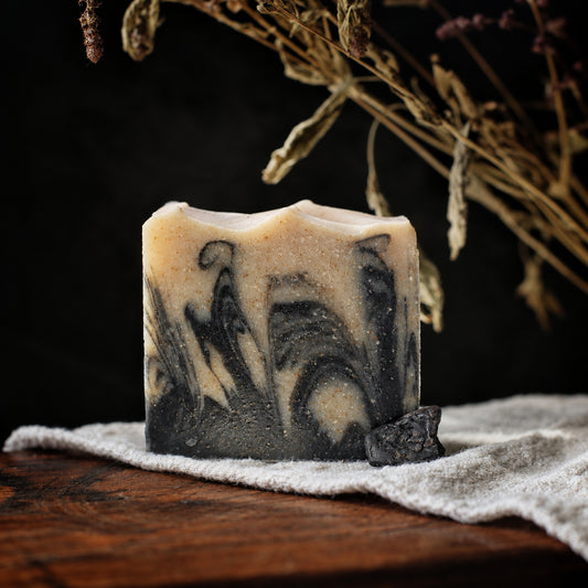 A handmade rectangular bar soap with a swirled black, gray, and cream pattern, resting on a white towel over a wooden surface with dramatic sunlight and dried flowers in the background.