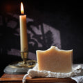 A rectangular bar of pale beige handmade soap with a rustic wavy top, resting on a piece of white lace over a wooden box next to a lit cream-colored candle in a brass holder against a dark background with soft shadows.