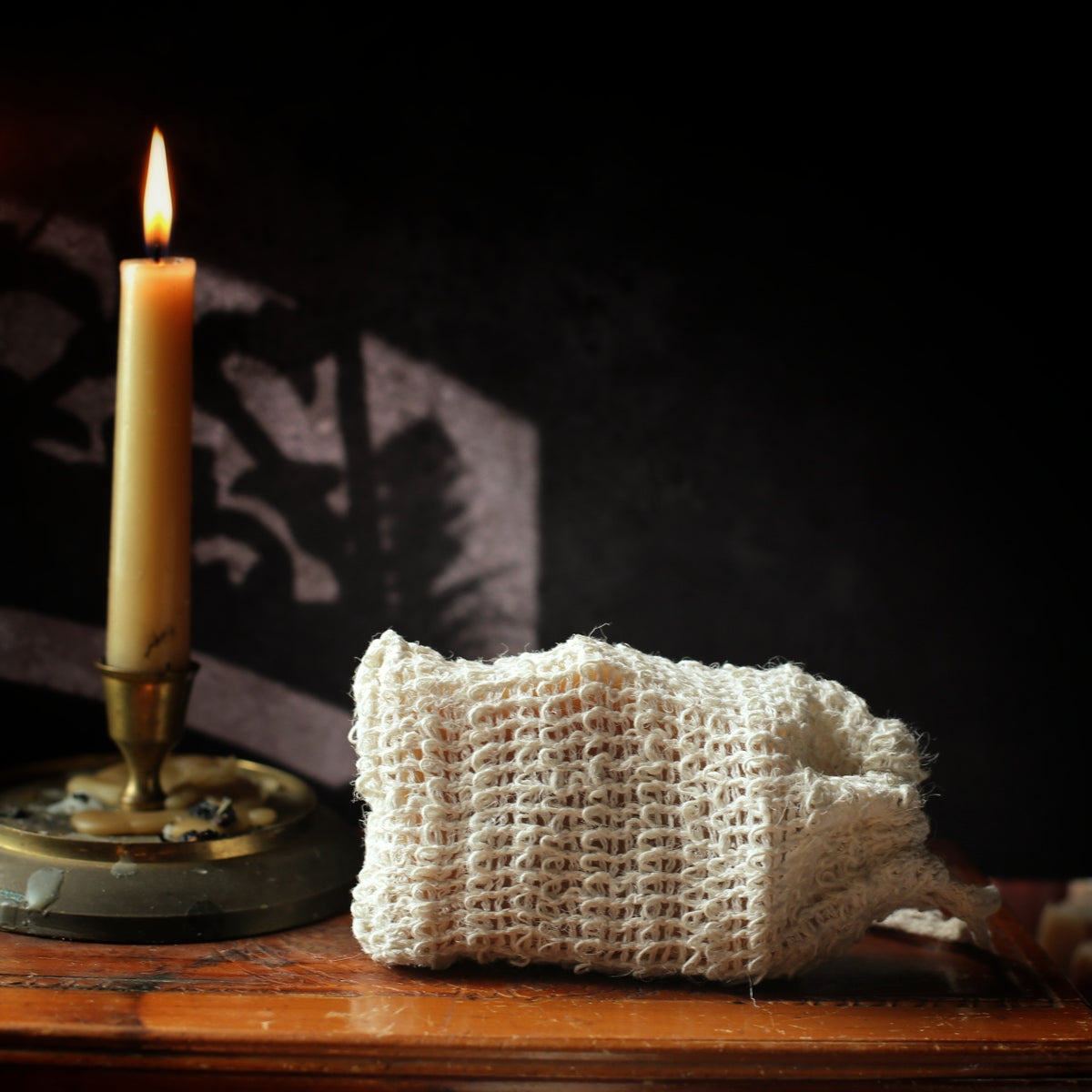 A small off-white Sisal pouch sits on a wooden surface next to a lit cream candle in a brass holder against a dark background with dramatic shadows.