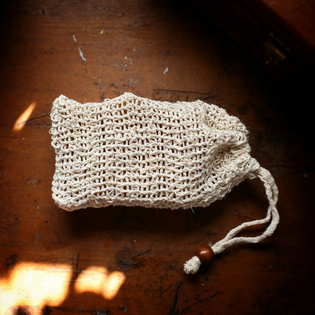 A cream-colored, knitted sisal drawstring pouch with a wooden bead accent, resting on a dark rustic wooden surface.