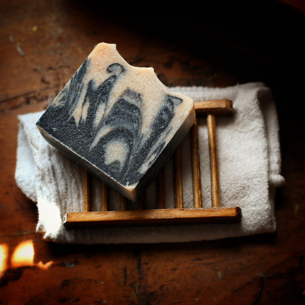 A rectangular handmade soap bar with a dark gray and beige marbled swirl pattern, resting on a small wooden soap dish over a white towel on a rustic wooden table.