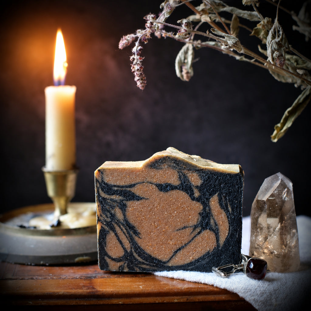 A styled product photo of a rectangular bar of soap with a dark and light brown marbled pattern, displayed on a wooden trunk next to a lit candle, a clear crystal point, and dried botanicals.